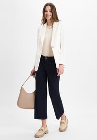 Blazer bianco, top beige, jeans a gamba larga blu scuro, mocassini spessi chiari e una borsa beige con un design curvo.