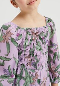 Junges Mädchen trägt ein lila Kleid mit grünen und pinken Blumenmustern sowie Raffung am Oberteil und Puffärmeln.