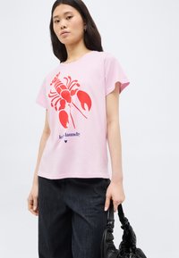 Νεαρή γυναίκα που φορά ροζ μπλούζα με κόκκινο γραφικό αστακού και κείμενο, μαύρο παντελόνι, κρατώντας μια μαύρη τσάντα με ρυθμιζόμενο σούρωμα.