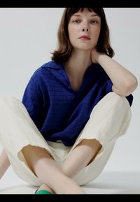 Femme aux cheveux bruns courts, vêtue d'une chemise bleue à motifs et d'un pantalon crème, assise jambes croisées sur un fond blanc, reposant sa main sur son cou.