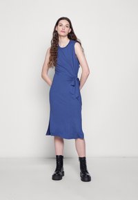 Vestido azul sem mangas com um detalhe amarrado na cintura, até ao joelho, feito de um tecido suave. Usado com botas pretas robustas. Fundo liso.