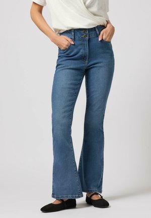 Femme portant un jean évasé taille haute bleu, un T-shirt blanc, des ballerines noires, debout les mains dans les poches devant un fond uni.