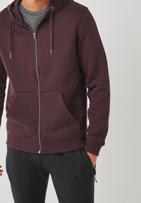 Sudadera con cremallera en burdeos, hecha de tela suave, con bolsillos frontales, capucha con cordón y puños y hem en canalé.