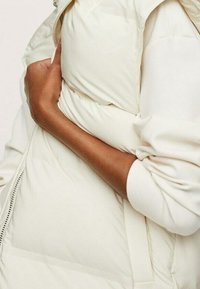 Gilet matelassé crémeux avec un col montant, présentant une finition lisse. Coutures détaillées et fermeture éclair sur un côté.