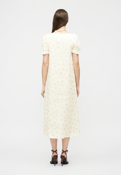 Γυναίκα με μακριά καστανά μαλλιά που φοράει λευκό midi φόρεμα με φουσκωτά μανίκια και κίτρινο λουλουδάτο μοτίβο, συνδυασμένο με μαύρα σανδάλια με ψηλό τακούνι, γυρισμένη πλάτη.