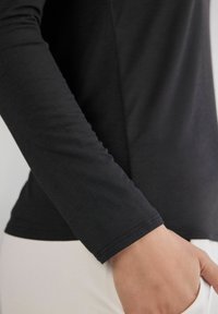 Schwarzes Langarmshirt mit glatter Textur und figurbetontem Schnitt. Verfügt über einen einfachen, genähten Saum und eine schlanke Silhouette, die am Handgelenk sichtbar ist.