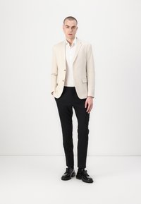 Lichtbeige blazer met een gestructureerde afwerking, wit overhemd met knopen, zwarte slim-fit broek en zwarte veterschoenen. Model staat rechtop.