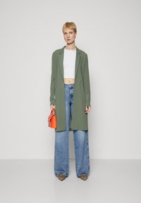 Sacou lung verde peste un top alb scurt și blugi largi de culoare albastru deschis. Accesoriile includ o geantă portocalie și pantofi cu vârf ascuțit.