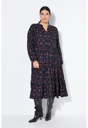 Kvinde iført en sort midi-kjole med pink hjerter og sløjfemønstre, lange ærmer, kombineret med sorte knæhøje støvler og øreringe med ringe.