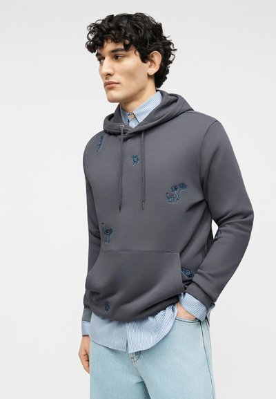 Νεαρός με σγουρά μαλλιά που φοράει γκρι φούτερ με κεντήματα πάνω από ένα μπλε ριγέ πουκάμισο και ανοιχτόχρωμο τζιν, στέκεται με τα χέρια στις τσέπες.