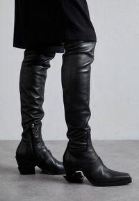 Black leather over-the-knee boots with a slouchy design, square toe, and a block heel. Features a side zipper and ankle strap hardware.