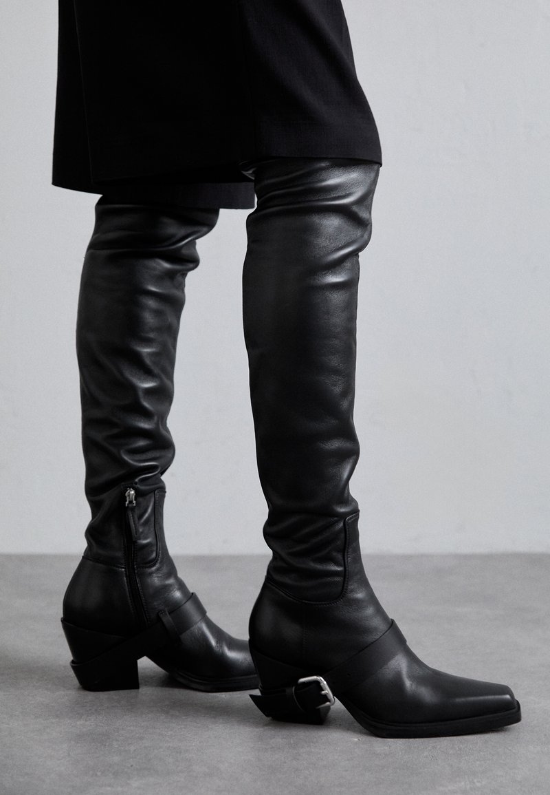 Black leather over-the-knee boots with a slouchy design, square toe, and a block heel. Features a side zipper and ankle strap hardware.