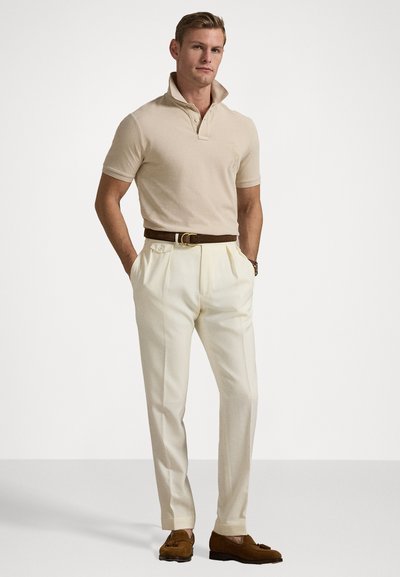 Beige lyhyet hihaton polo, jossa on kohotettu kaulus, kermaiset pituuslahkeiset housut, ruskea vyö ja ruskeat samettiset loafersit. Yksinkertainen muotoilu, teksturoitu kangas.