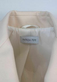 Interior de una prenda blanca con una textura de satén suave, con una etiqueta que dice "PATRIZIA PEPE" y un lazo de tela para colgar.