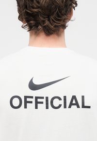 Baltas medvilninis marškinėlis su juodu „Nike“ logotipu ir ryškiu užrašu „OFFICIAL“ nugaroje. Trumpomis rankovėmis, apvalia iškirpte ir lygiu paviršiumi.