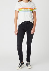 Camiseta blanca con el logo "Wrangler" multicolor, jeans ajustados negros y zapatillas negras con suelas blancas, sobre un fondo neutro.