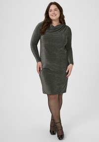 Femme souriante dans une robe noire pailletée de longueur genou avec des manches longues, des collants noirs transparents et des chaussures Mary Jane noires en vernis.