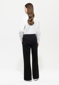 Weißes langärmeliges Hemd mit Knöpfen und glatter Textur, kombiniert mit hochtaillierten, weit geschnittenen schwarzen Hosen, die zwei Gesäßtaschen zeigen.