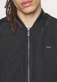 Černá bomber bunda s hladkou texturou, stříbrným zipem, žebrovaným límcem a malým logem Calvin Klein na hrudi.
