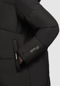 Doudoune noire avec une texture lisse, un design matelassé, une poche latérale et une étiquette de marque sur la manche avec des détails de code-barres.