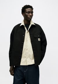 Άνδρας που φοράει μαύρο μπουφάν με στρώσεις πάνω από μπεζ πουκάμισο και σκούρο τζιν, στέκεται μπροστά σε απλό λευκό φόντο, κοιτάζοντας προς το πλάι.