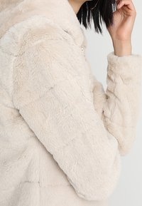 Beige faux fur jakke med en fyldig tekstur, lynlås-lukning og en afslappet pasform. Har en krave og quiltet mønster på stoffet.