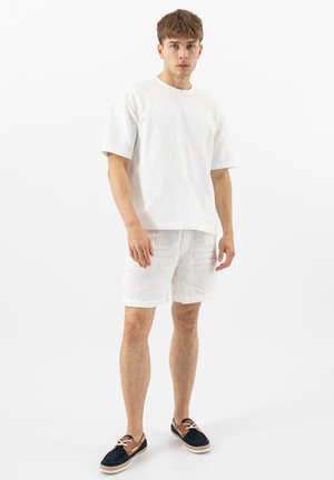 Mladý muž v jednoduchém bílém tričku s krátkým rukávem a bílých šortkách, stojící s uvolněnýma rukama, obutý v námořnicky modrých a hnědých šněrovacích botách.