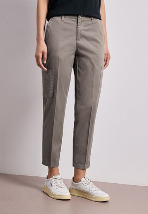 Personne portant un pantalon cargo gris court avec des poches latérales et des baskets blanches, debout devant un fond clair uni.