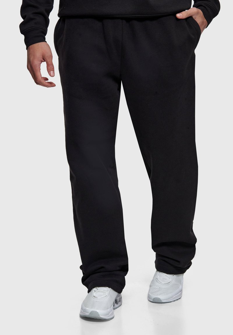 Homme portant un pantalon de survêtement noir et des baskets blanches, debout les mains dans les poches devant un fond uni.