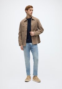 Μπεζ πουκάμισο-ζακέτα από μείγμα μαλλιού με δύο μπροστινές τσέπες, φορεμένη πάνω από ένα μπλε σκούρο πουκάμισο, συνδυασμένη με ανοιχτό μπλε τζιν και μπεζ αθλητικά παπούτσια.