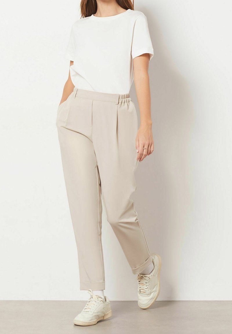 Pantalon plissé beige avec taille élastique, poches latérales et jambes fuselées. Associé à un t-shirt blanc à manches courtes et des baskets beige.