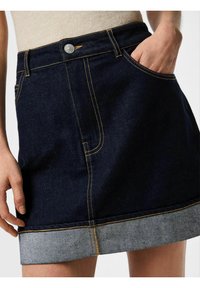 Gonna di denim blu scuro con orlo grezzo blu chiaro. Presenta cuciture gialle, una tasca frontale e una chiusura con bottone in vita.