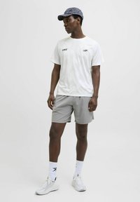 Hvid kortærmet skjorte med subtilt mønster, grå shorts, hvide sneakers og en mørk mønstret kasket. Enkel design med logo detaljer.