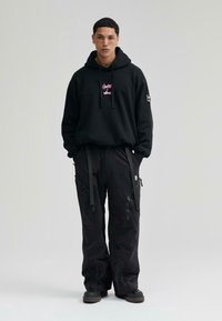 Sudadera negra con texto gráfico rosa, combinada con pantalones cargo negros de corte holgado que tienen bolsillos con cremallera y correas ajustables. Calzado negro.