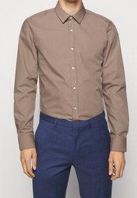Brown long-sleeve button-up shirt in smooth fabric, featuring a classic collar and subtle sheen, paired with navy textured trousers.