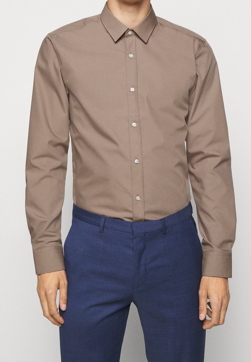 Brown long-sleeve button-up shirt in smooth fabric, featuring a classic collar and subtle sheen, paired with navy textured trousers.