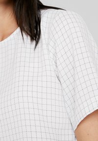 Biały top z krótkim rękawem z wzorem w kratę z małych czarnych kwadratów, o gładkiej fakturze i luźnym kroju.