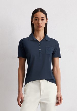 Marineblauwe poloshirt met korte mouwen, knoopsluiting en een borstzak. Zachte stof en getailleerd ontwerp. Gemaakt in combinatie met witte broek.