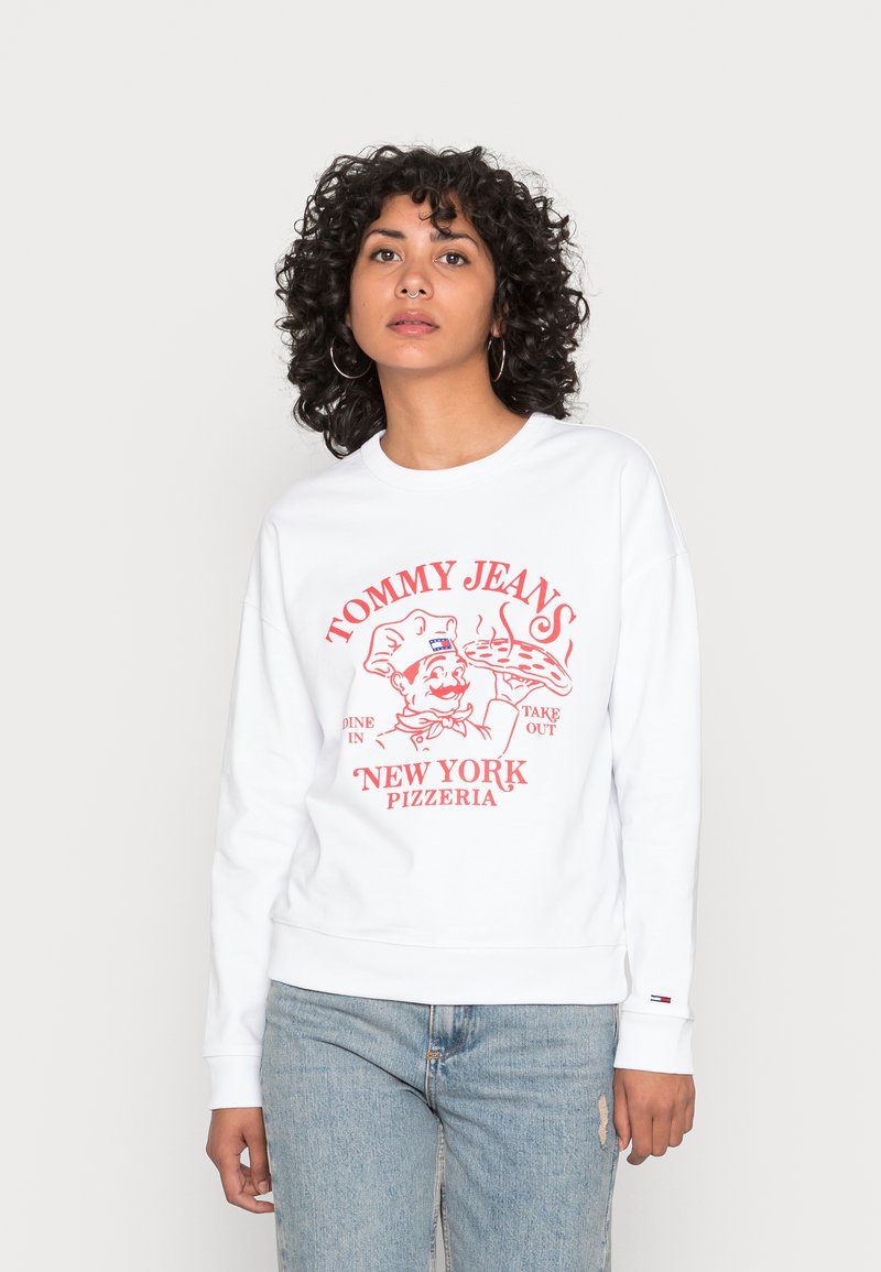 Biely sveter s červeným grafickým dizajnom, ktorý zobrazuje kuchára a text "TOMMY JEAN'S NEW YORK PIZZERIA." Mäkký materiál a rebrované manžety.