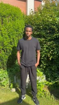 Svart kortärmad t-shirt, svarta joggers och svarta sneakers bärs av en stående figur i en frodig grön trädgård med solljus.