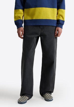 Personne portant un jean noir ample, un tee-shirt à manches longues rayé bleu et jaune, et des chaussures slip-on à carreaux noir et blanc, debout.