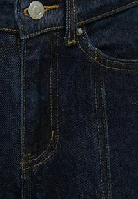 Tamno indigo traperice s žutim šavovima, opremljene srebrnom metalnom gumbom i prednjim džepom. Tekstura izgleda glatko i izdržljivo.