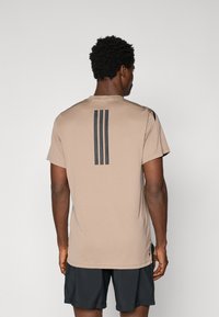 T-shirt de sport marron clair à manches courtes, le dos présente trois rayures verticales noires, fabriqué en matériau respirant avec une texture lisse.