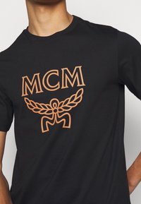 Camiseta de algodón negra con un logo impreso en naranja que dice "MCM" y un diseño de alas estilizadas. Mangas cortas, cuello redondo clásico.