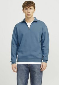 Blue zip-up sweatshirt made of soft fabric with a high collar and ribbed cuffs, paired with light blue jeans for a casual look.
