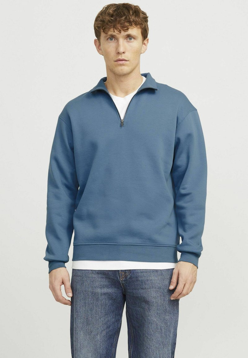 Blue zip-up sweatshirt made of soft fabric with a high collar and ribbed cuffs, paired with light blue jeans for a casual look.