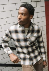 Camicia a quadri nei toni del nero, bianco e marrone; presenta bottoni, un colletto e una tasca sul petto; mostrata su uno sfondo piastrellato.