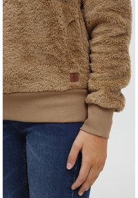 Tan fuzzy pullover lavet af blødt, tekstureret stof med ribbede manchetter og kant. Har et lille brunt mærke foran.