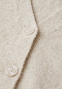 Bežový pletený cardigan s texturovaným povrchem, se třemi bílými knoflíky a designem s výstřihem do V. Detailní záběr zdůrazňuje strukturu látky.