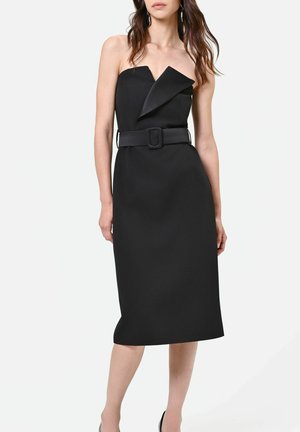 Robe midi noire bustier avec détail d'encolure asymétrique et large ceinture à boucle rectangulaire portée par une femme debout.